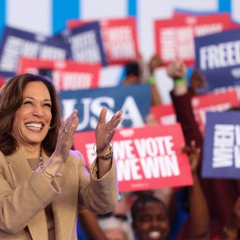 Kamala Harris claps; people behind her hold up signs in support.