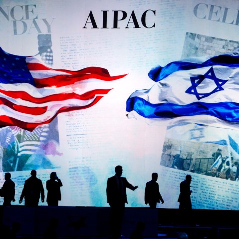 People are silhouetted before a screen that reads "AIPAC" and shows a visual of the American flag and the Israeli flag.