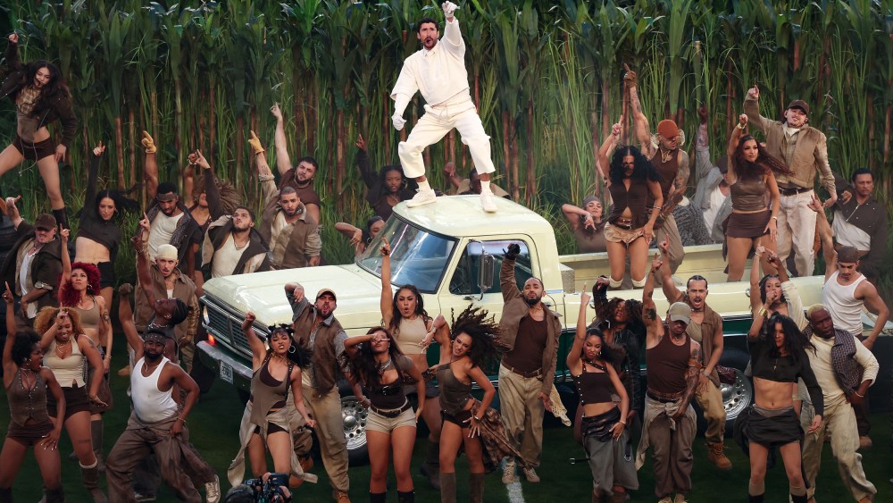 Bad Bunny stands on the roof of a light yellow truck. People dance in the truck bed and on the football field below.