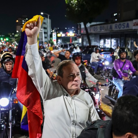 Juan Pablo Guanipa, center, stands in the street, holding a Venezuelan flag up with one hand. He is surrounded by people on motorcycles.