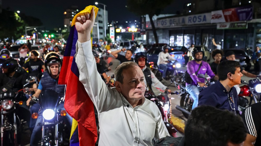 Juan Pablo Guanipa, center, stands in the street, holding a Venezuelan flag up with one hand. He is surrounded by people on motorcycles.