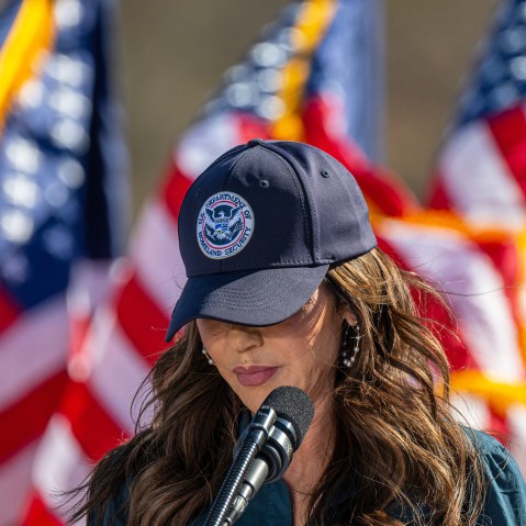 Kristi Noem with her head tilted down, her "U.S. Department of Homeland Security" baseball cap obscuring her eyes.