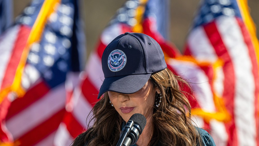 Kristi Noem with her head tilted down, her "U.S. Department of Homeland Security" baseball cap obscuring her eyes.