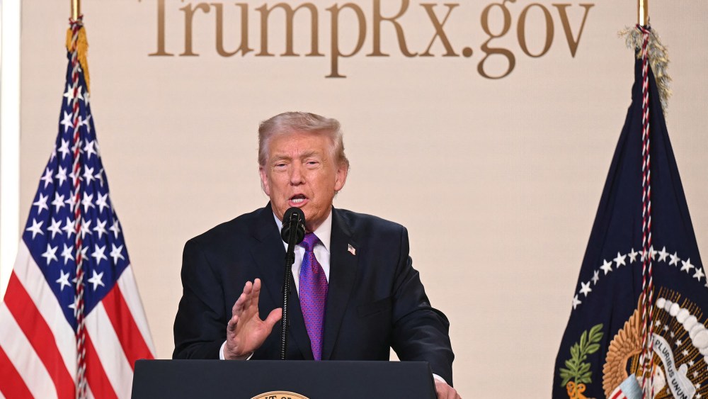 Donald Trump speaks into a microphone at a podium. The website "TrumpRx.gov" is on the wall behind him.