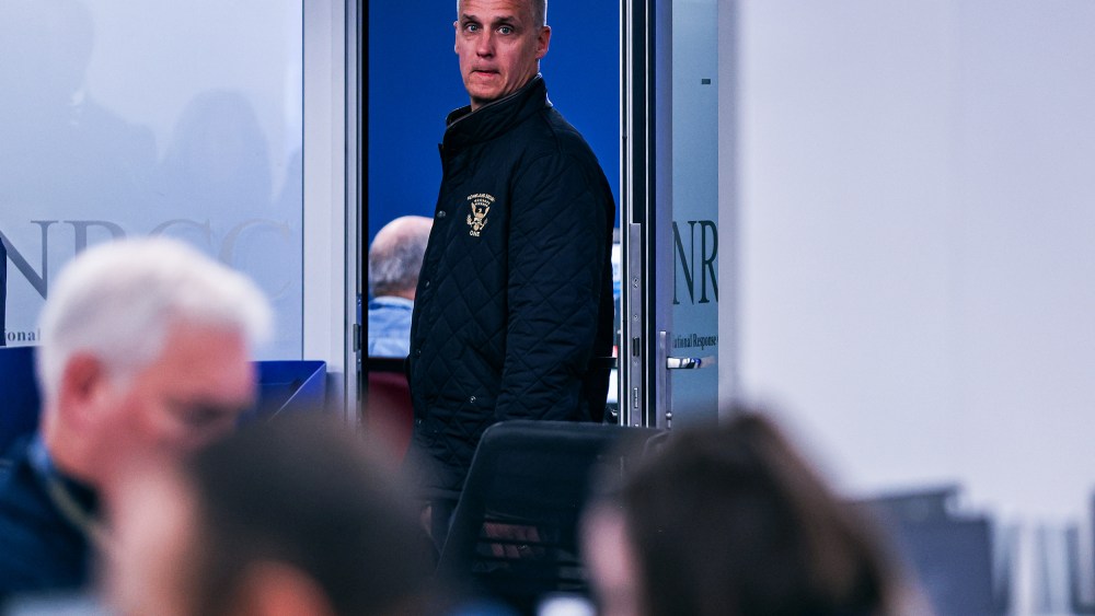 Corey Lewandowski stands in a doorway. Blurred in the foreground, people work at their desks.