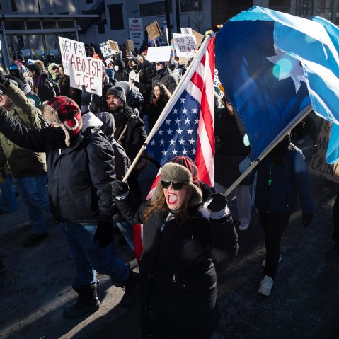 A female protestor shouts as she carries a flag in the foreground where others follow her during a daytime demonstration.