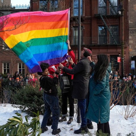 People raise a rainbow flag on a pole in a park. A crowd is gathered behind, watching and filming.