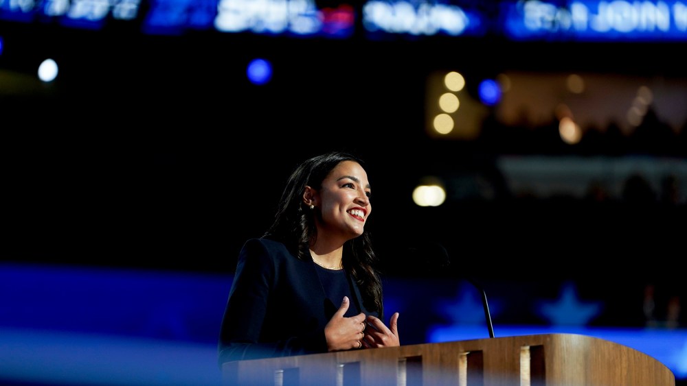 Alexandria Ocasio-Cortez smiles while standing behind a podium.