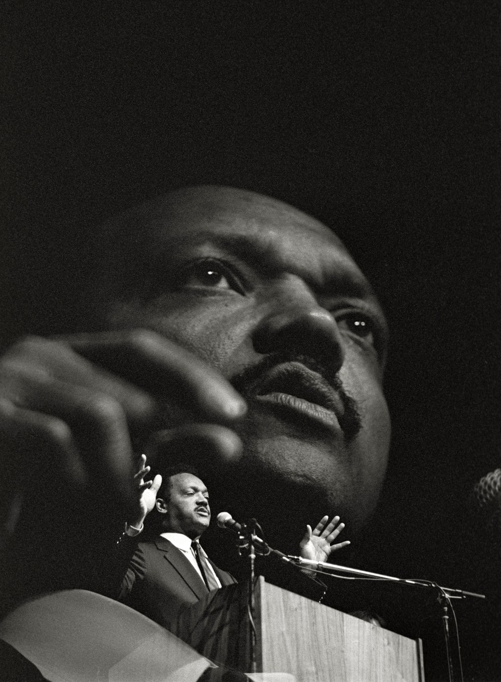 A vertical, black-and-white double exposure photo of Rev. Jesse Jackson. The first layer is a medium frame of him speaking into a microphone at a podium; the second layer is a close up of him speaking into the mic.