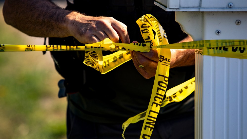 The hands of a police office tie yellow tape that says "Police Line Do Not Cross."