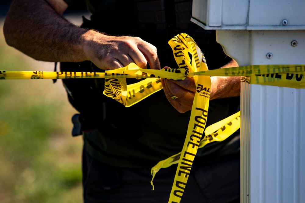 The hands of a police office tie yellow tape that says "Police Line Do Not Cross."