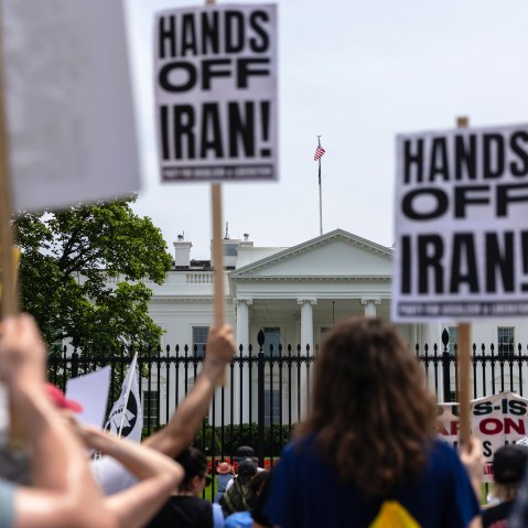 Out of focus in the foreground, people hold picket signs that read "Hands Off Iran!" In focus in the background is the White House.