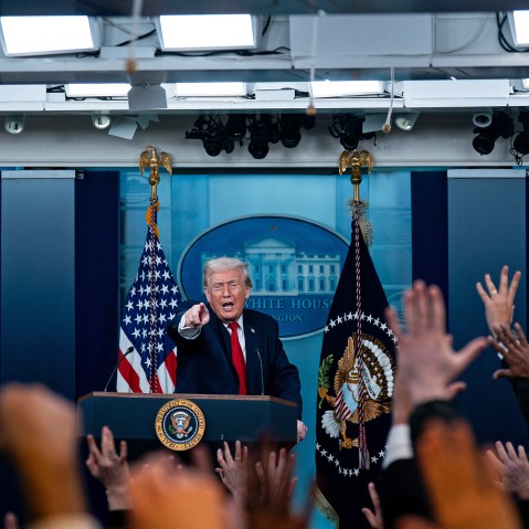 Donald Trump, in focus, stands a podium and points in front of him. Out of focus in the foreground, many people raise their hand.