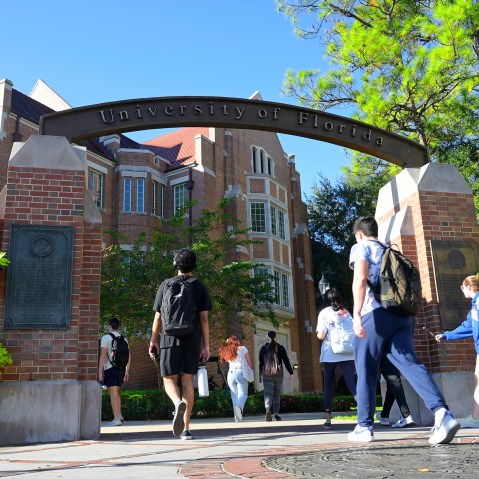 Students walk under the arch of University of Florida entrance on a sunny day.