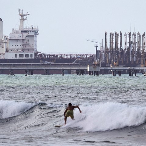 A man is seen from behind as he surfs in front of an oil tanker in the distance.