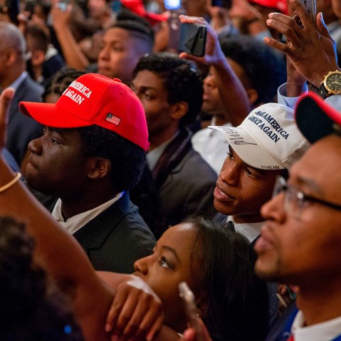 Audience members, most of them Black, wear MAGA hats and hold up their phones to take photos.