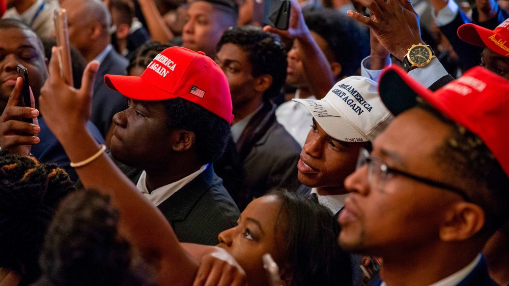 Audience members, most of them Black, wear MAGA hats and hold up their phones to take photos.