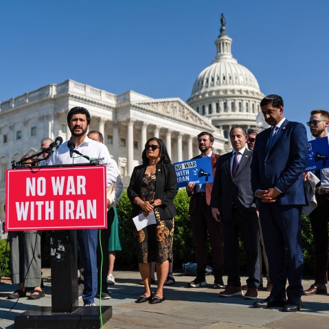 Greg Casar stands behind a podium that has a red sign that says "NO WAR WITH IRAN." He is surrounded by other people, and in the background is the Capitol.
