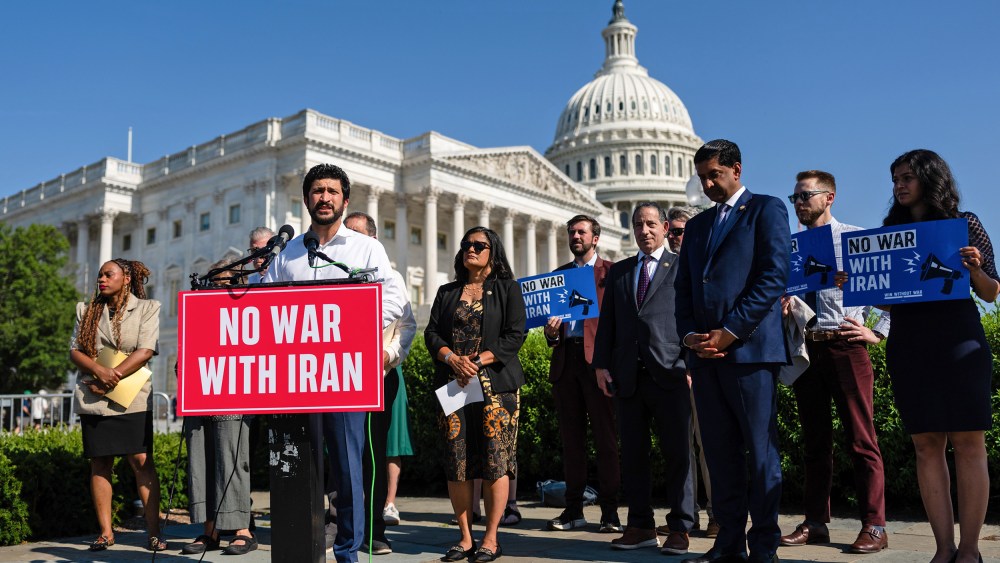 Greg Casar stands behind a podium that has a red sign that says "NO WAR WITH IRAN." He is surrounded by other people, and in the background is the Capitol.