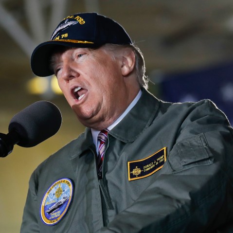 Donald Trump speaks into a microphone while wearing a USS Gerald R. Ford jacket and hat.