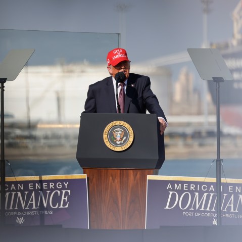 Donald Trump speaks into a microphone at a podium. He's wearing a red hat that says "Gulf of America."