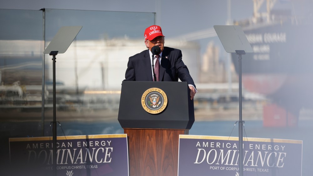 Donald Trump speaks into a microphone at a podium. He's wearing a red hat that says "Gulf of America."