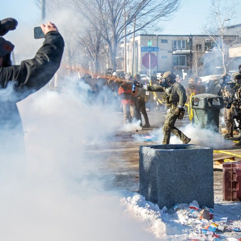 A person on the left films while a federal agent, center, sprays an orange substance ahead. On the right, other agents standby.