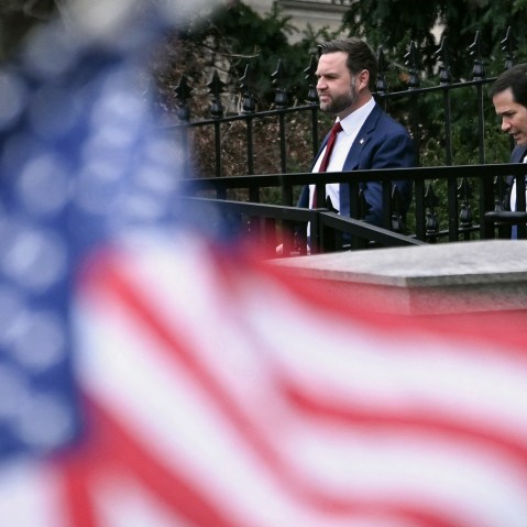 Out of focus in the foreground is the American flag, occupying the left and bottom of the frame. JD Vance, left, and Marco Rubio are in focus in the background, seen walking out of a building.