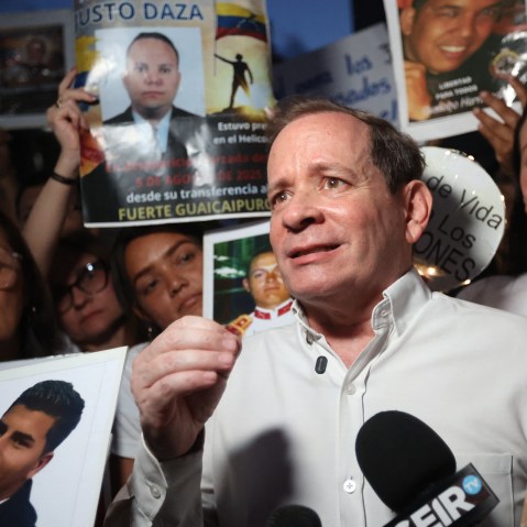 Juan Pablo Guanipa stands in front of a microphone with a crowd of people behind him holding signs for the release of other Venezuelan prisoners.