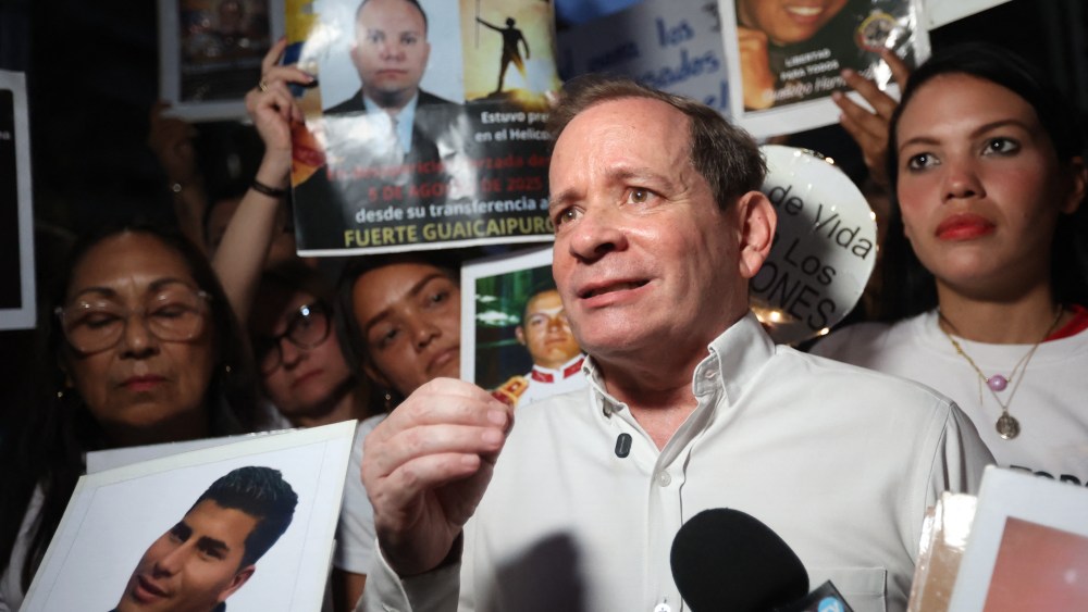 Juan Pablo Guanipa stands in front of a microphone with a crowd of people behind him holding signs for the release of other Venezuelan prisoners.