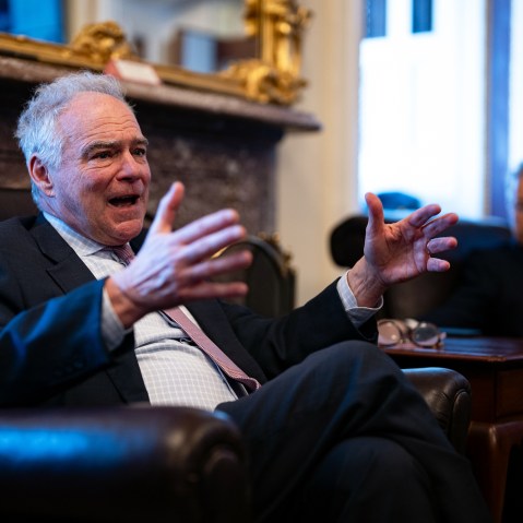 Tim Kaine, in focus on the left, gestures with his hands, while Rand Paul, out of focus on the right, looks at him.