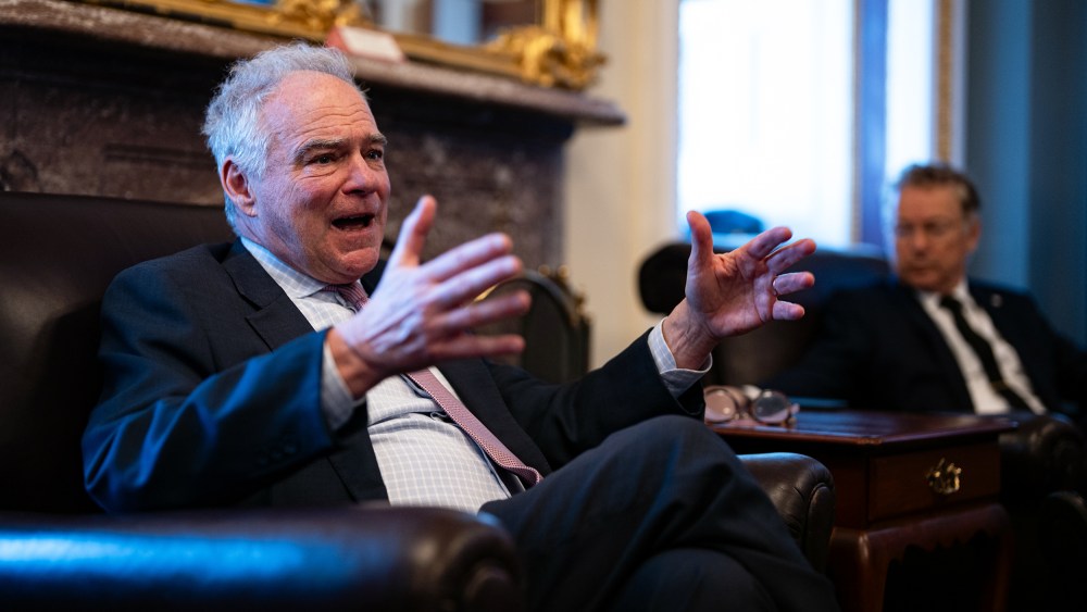 Tim Kaine, in focus on the left, gestures with his hands, while Rand Paul, out of focus on the right, looks at him.
