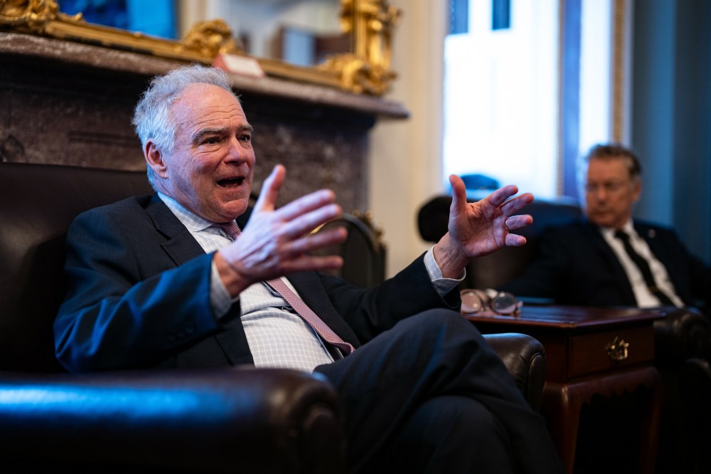 Tim Kaine, in focus on the left, gestures with his hands, while Rand Paul, out of focus on the right, looks at him.