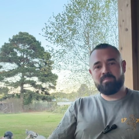 Aaron Spencer sits at a porch as he speaks. Turkeys can be seen in the background.