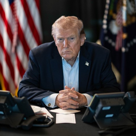 Donald Trump sits at a table with two phones in front of him and U.S. flags behind him.
