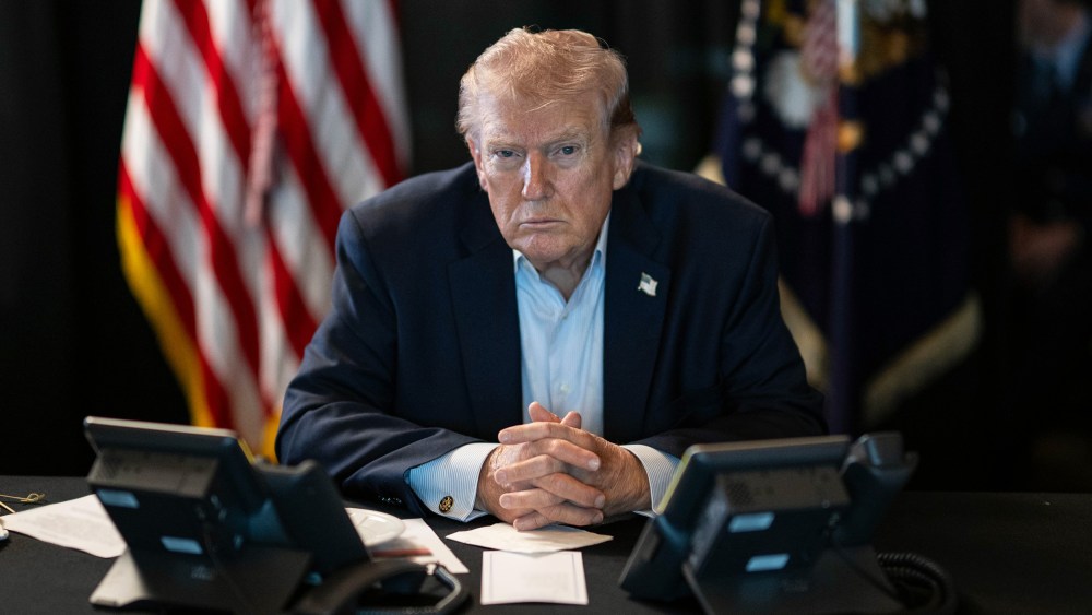 Donald Trump sits at a table with two phones in front of him and U.S. flags behind him.