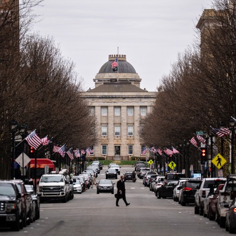 A street lined with cars leads up to the North Carolina State Capitol. A person crosses in the middle of the street, and American flags are on each of the lampposts.
