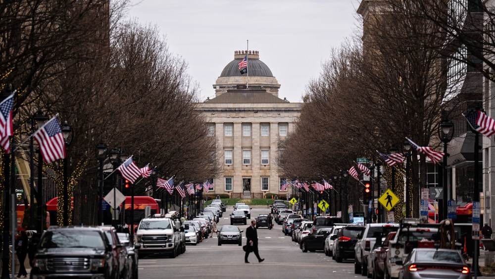 A street lined with cars leads up to the North Carolina State Capitol. A person crosses in the middle of the street, and American flags are on each of the lampposts.