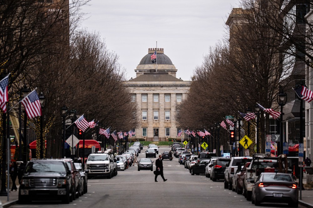 A street lined with cars leads up to the North Carolina State Capitol. A person crosses in the middle of the street, and American flags are on each of the lampposts.