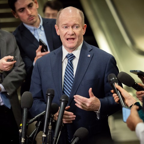 Chris Coons speaks while surrounded by reporters holding phones and microphones.