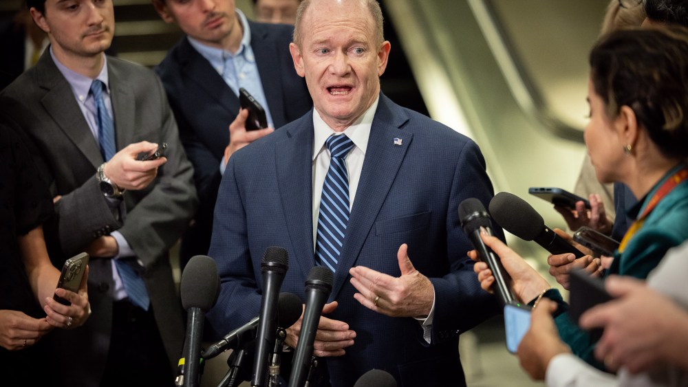 Chris Coons speaks while surrounded by reporters holding phones and microphones.