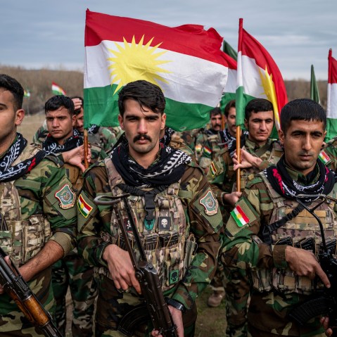 People in military uniforms holding guns and Kurdish flags stand in formation.