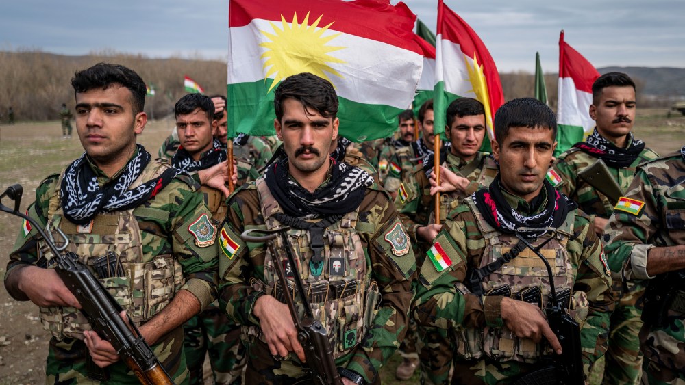 People in military uniforms holding guns and Kurdish flags stand in formation.