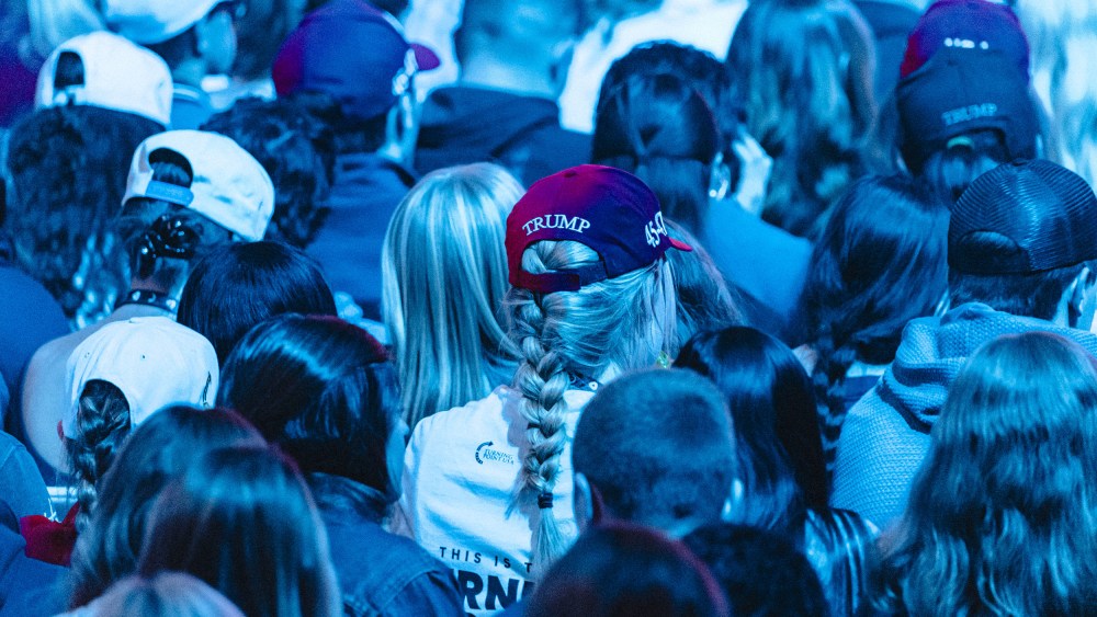 Backs of people's heads sitting in an audience. Multiple people wear Trump hats. The entire photo is tinted blue by the lights.