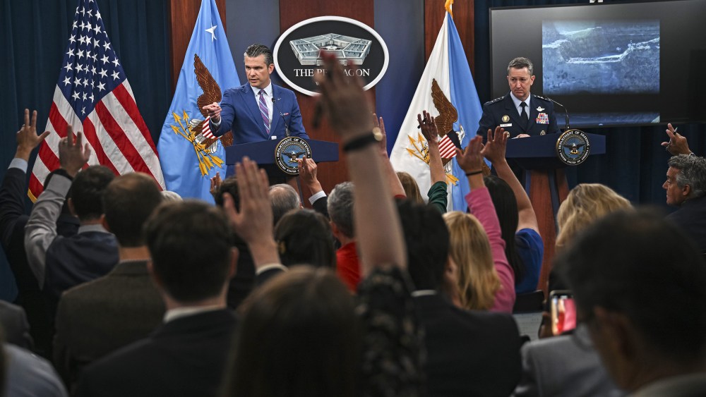 People in the foreground raise their hands; in focus in the background, Pete Hegseth, left, and Dan Caine stand behind podiums.