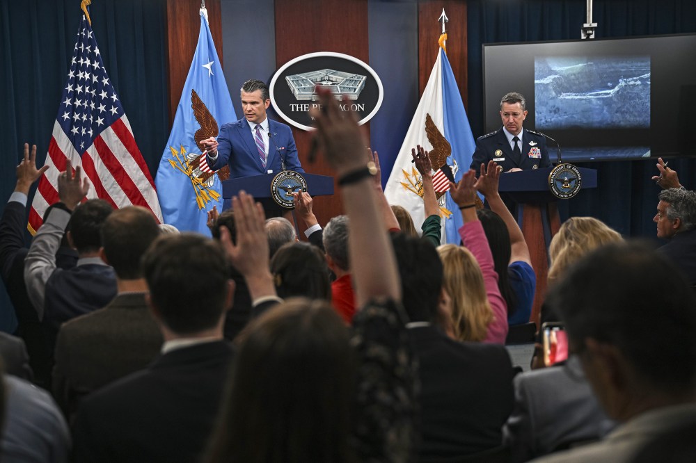 People in the foreground raise their hands; in focus in the background, Pete Hegseth, left, and Dan Caine stand behind podiums.