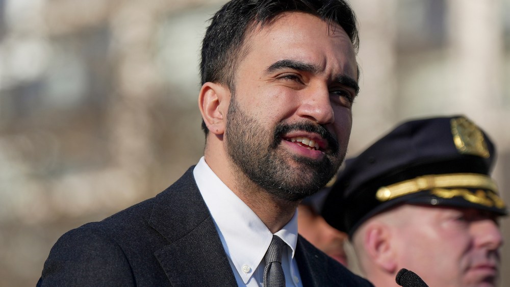 In this tight shot, Zohran Mamdani is seen speaking in front of a mic while a law enforcement officer stand behind to the right.