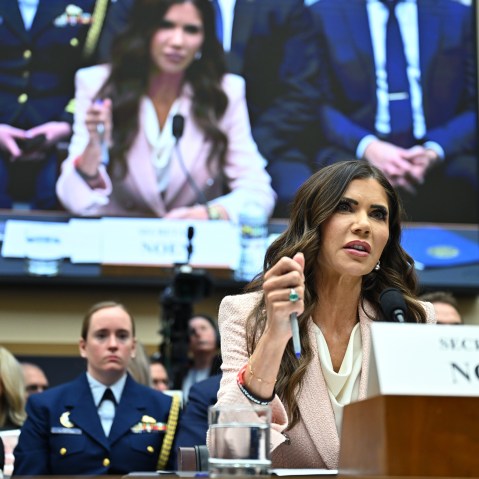 Kristi Noem holds a pen as she testifies where her image is also show on a screen in the background.