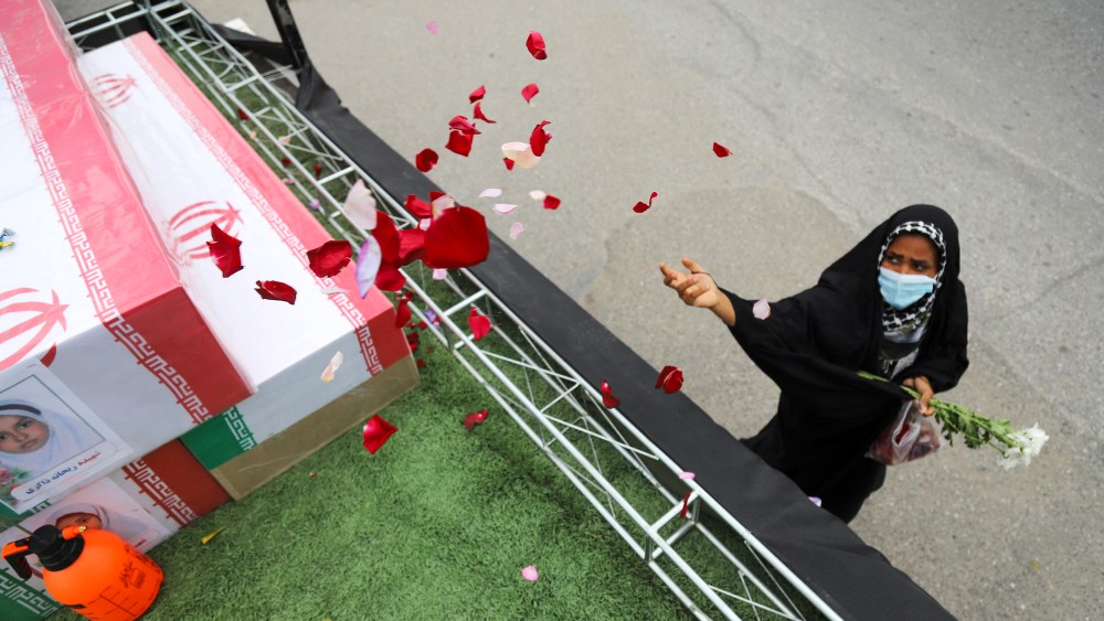 A woman is seen wearing a black hijab on the right side of the frame as she throws flower petals on a coffin.