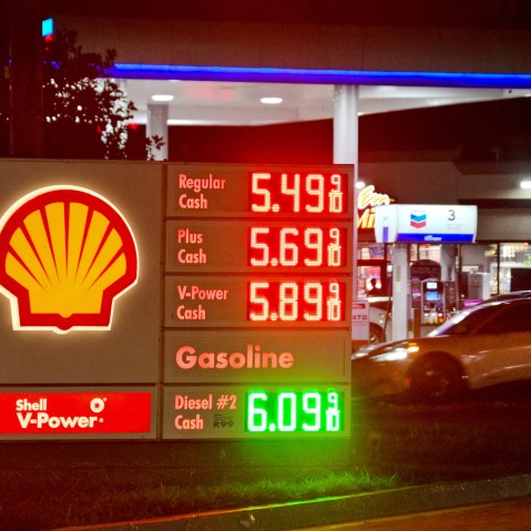 A gas station sign on the ground shows fuel prices while a car pulls into to get gas.